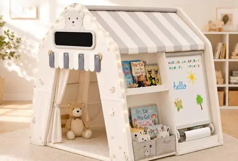 Casita de juego con estante de cuentos, canastas, pizarra blanca con rollo de papel y un oso de peluche en la entrada