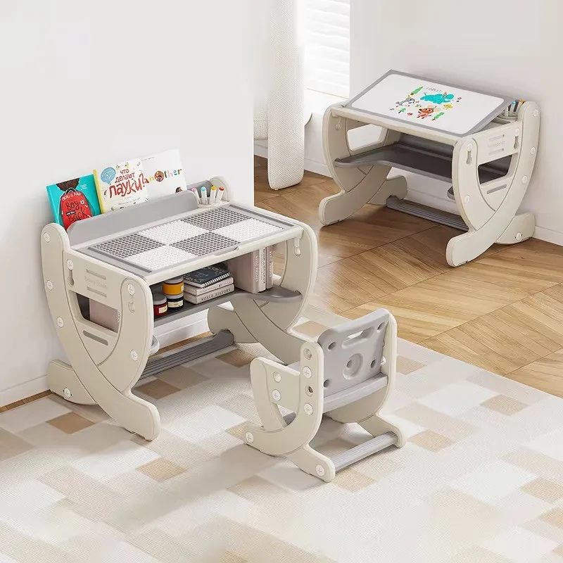 Escritorio infantil beige con tablero para armar bloques, cuentos atrás y silla a juego; al fondo otro escritorio con la pizarra levantada