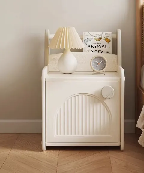 Mesita de noche junto a una cama, con una lámpara, un reloj y un cuento en el revistero