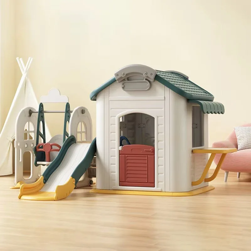 Casita de juego blanca con techo verde, puerta roja y mostrador amarillo, junto a un tobogán y un columpio