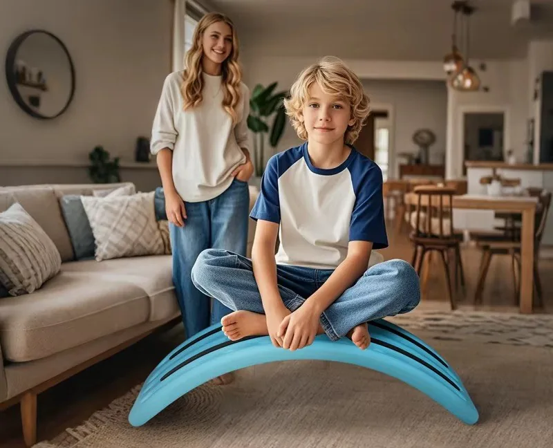 Niño sentado sobre la tabla de equilibrio volteada como puente, en una sala, con una mujer de pie detrás